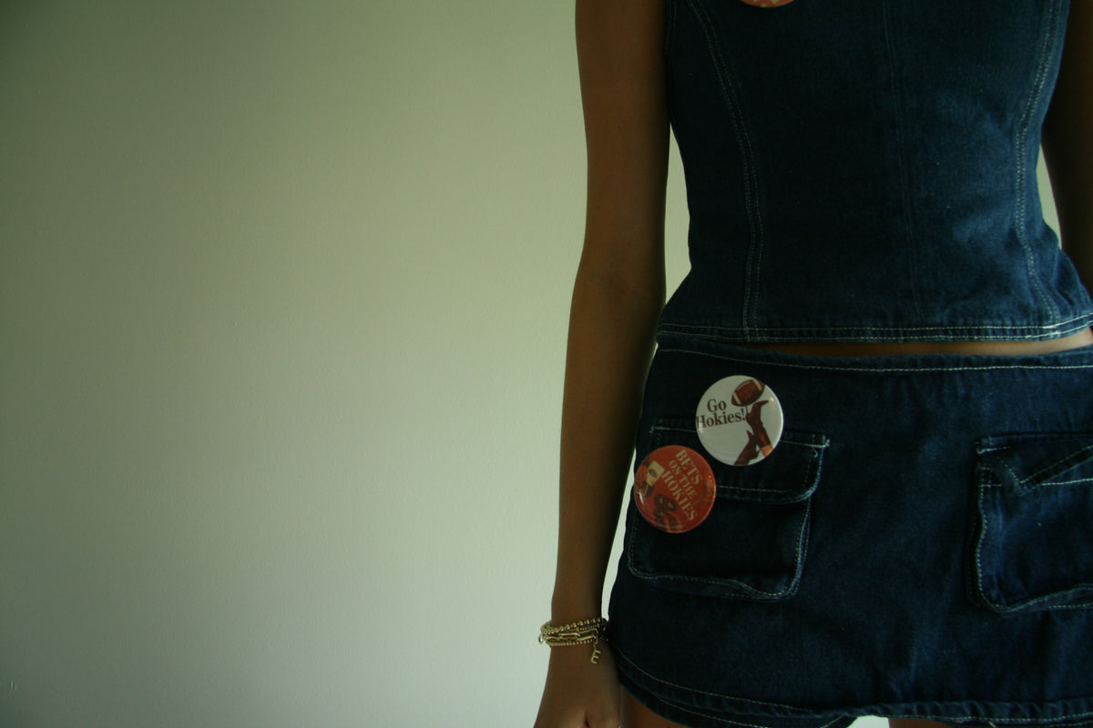 Close-up of a person wearing a dark blue denim dress or skirt with a sleeveless top, shown from the back. The denim garment has two back pockets, one of which holds two round pins, against a plain light background.